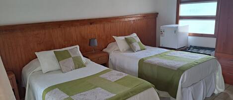 A hotel room with two beds, wooden headboard, bedside tables with lamps, a small refrigerator, and a window with blinds.