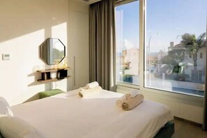 Habitación de hotel con un ventanal grande, una cama con toallas dobladas, un espejo y una estantería con artículos de aseo.