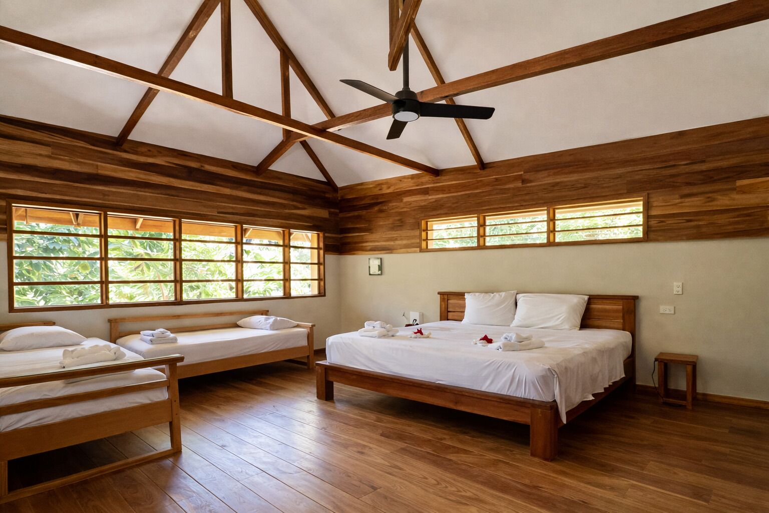 Una habitación con dos camas de madera, un ventilador de techo y grandes ventanas con vistas a zonas verdes.
