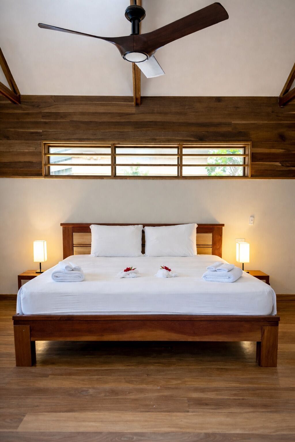 Una cama grande con ropa blanca, dos lámparas de noche y un ventilador de techo en una habitación con vigas de madera y una ventana con persianas horizontales.