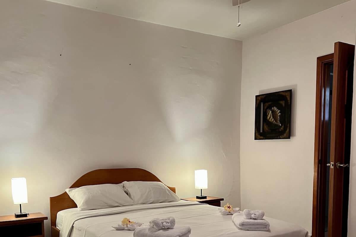 Una cama bien hecha con sábanas blancas, dos mesitas de noche con lámparas, un ventilador de techo y un cuadro en la pared.