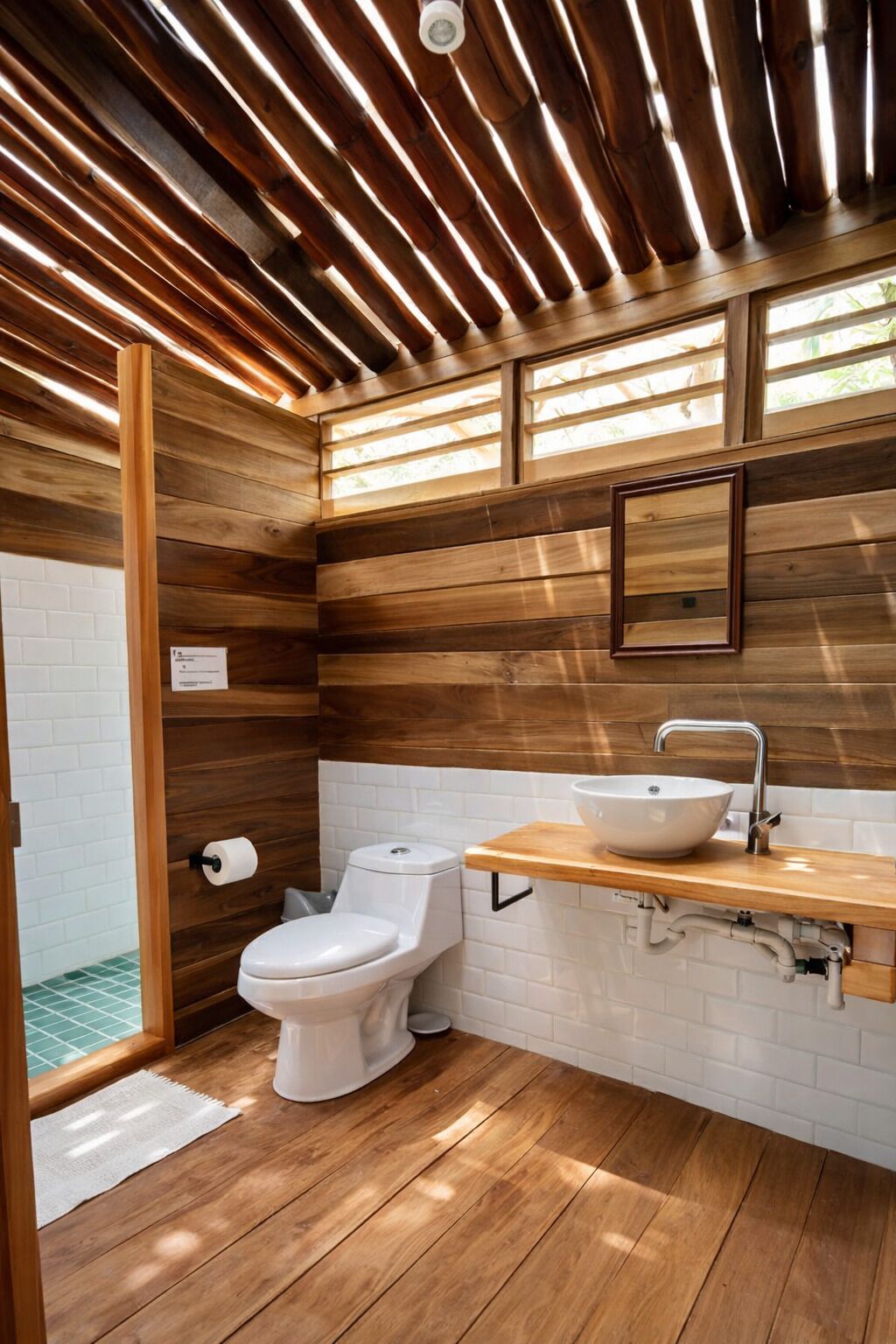 Un baño con paredes de madera, un inodoro blanco, un lavabo con espejo y una ventana sobre el lavabo.