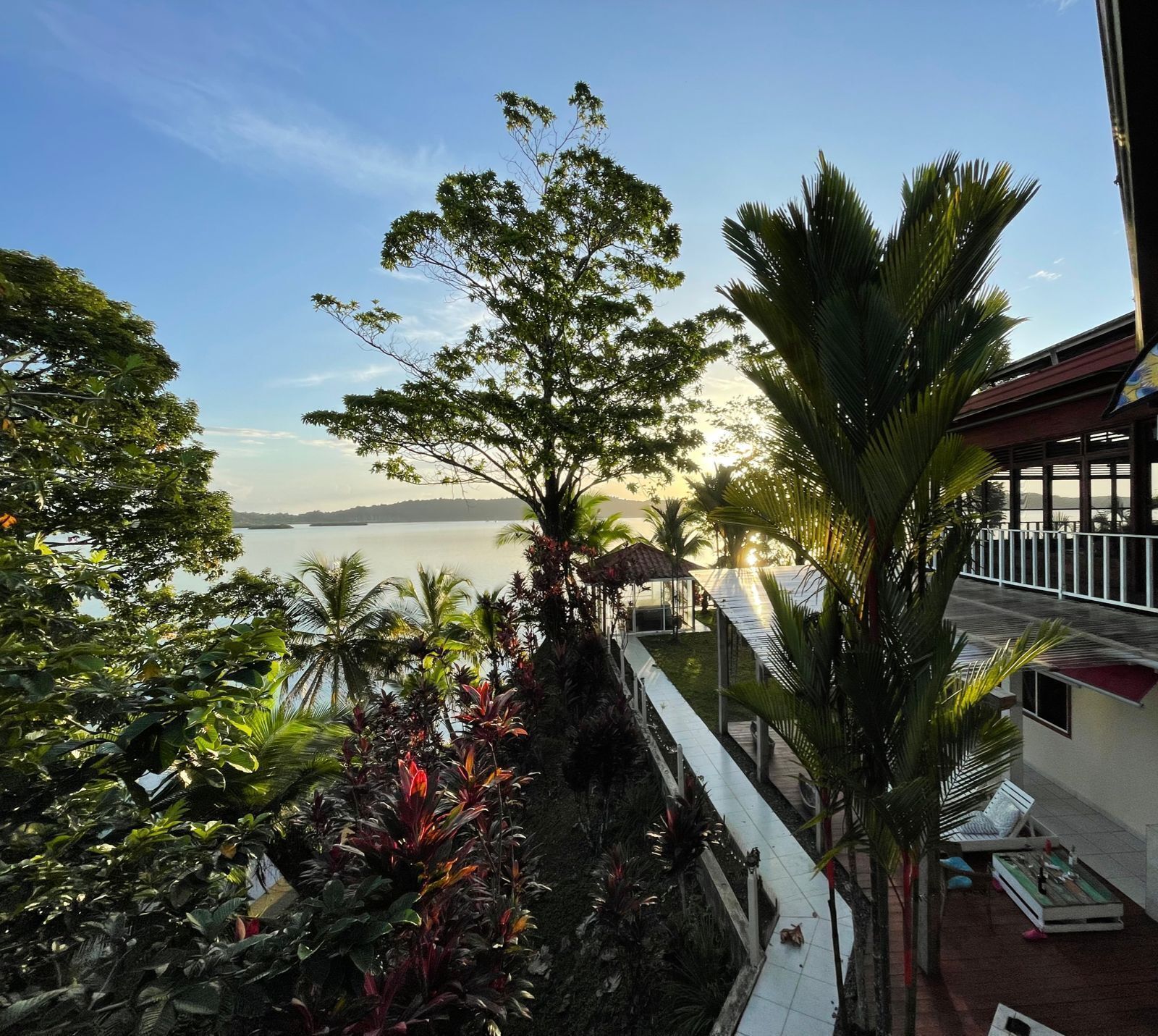 Un balcon offrant une vue sur une étendue d’eau paisible et sur des montagnes lointaines, le tout entouré d’une végétation tropicale luxuriante.