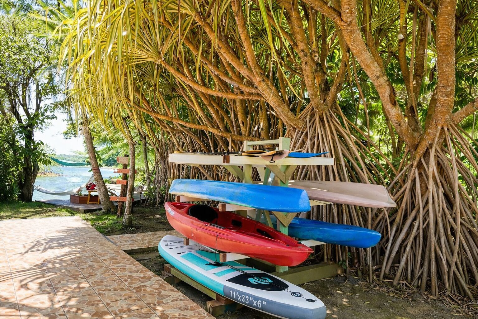 A row of kayaks and a paddleboard under a tree, with a tiled walkway and a hammock nearby.