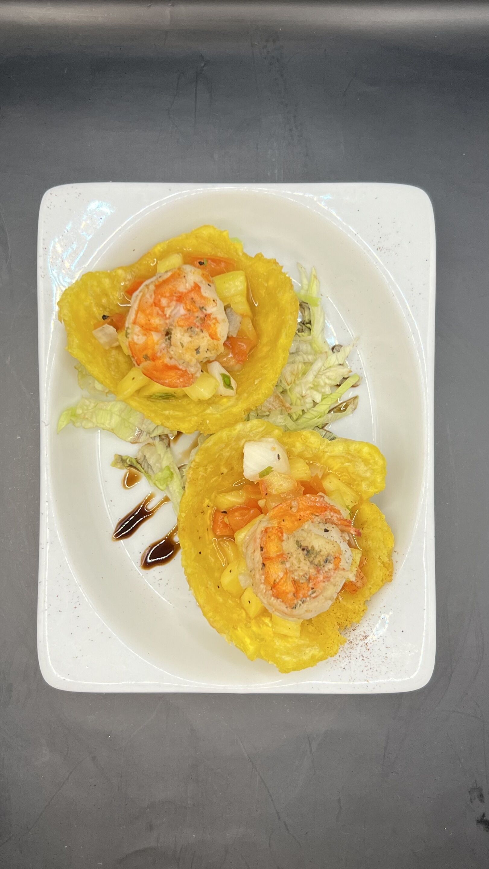 A white plate with two yellow-colored food items, possibly tacos, with a sauce and shredded lettuce on a metallic surface.