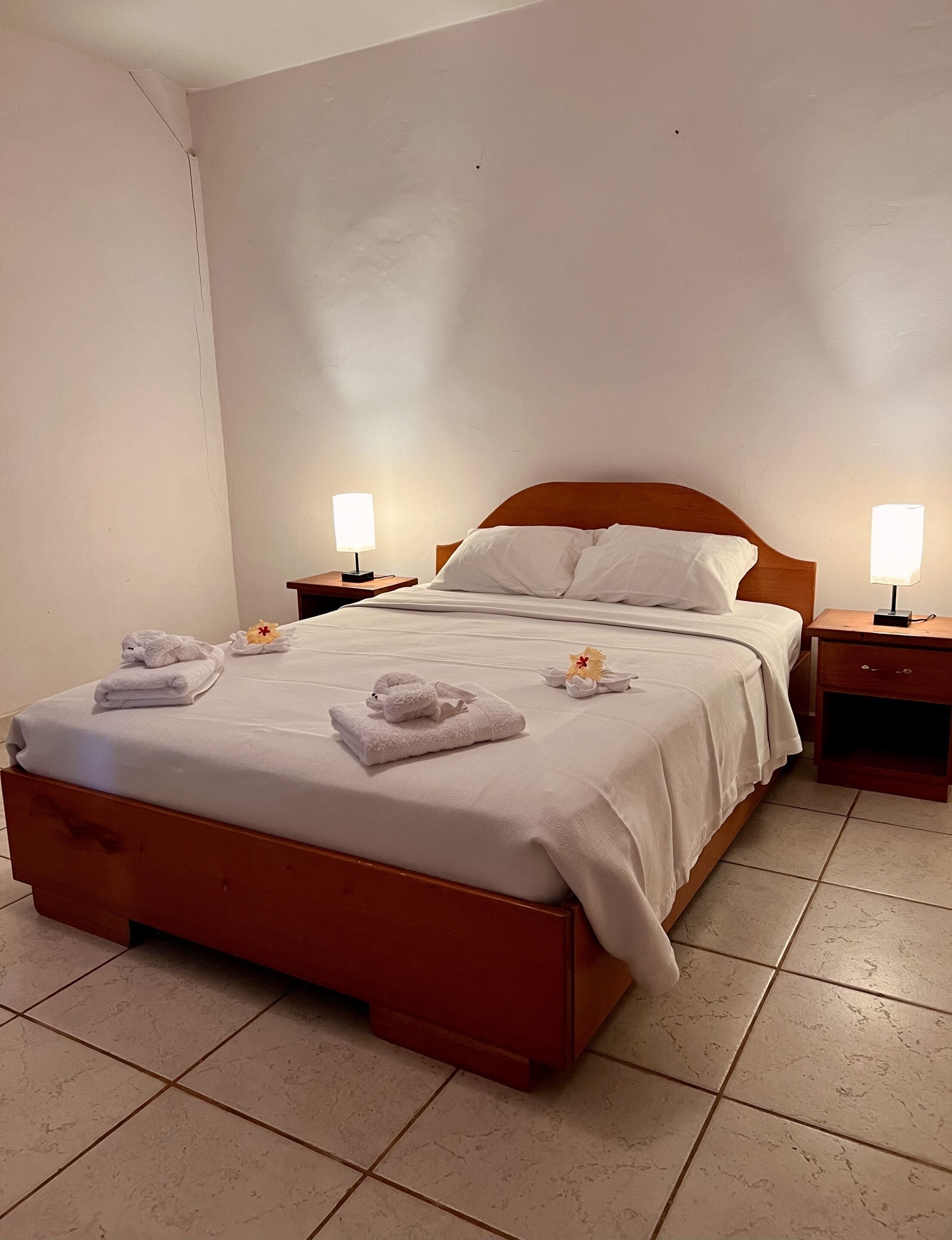 Ein ordentlich bezogenes Bett mit weißen Leinen und Handtüchern, zwei Nachttischlampen und ein Holz-Kopfteil.