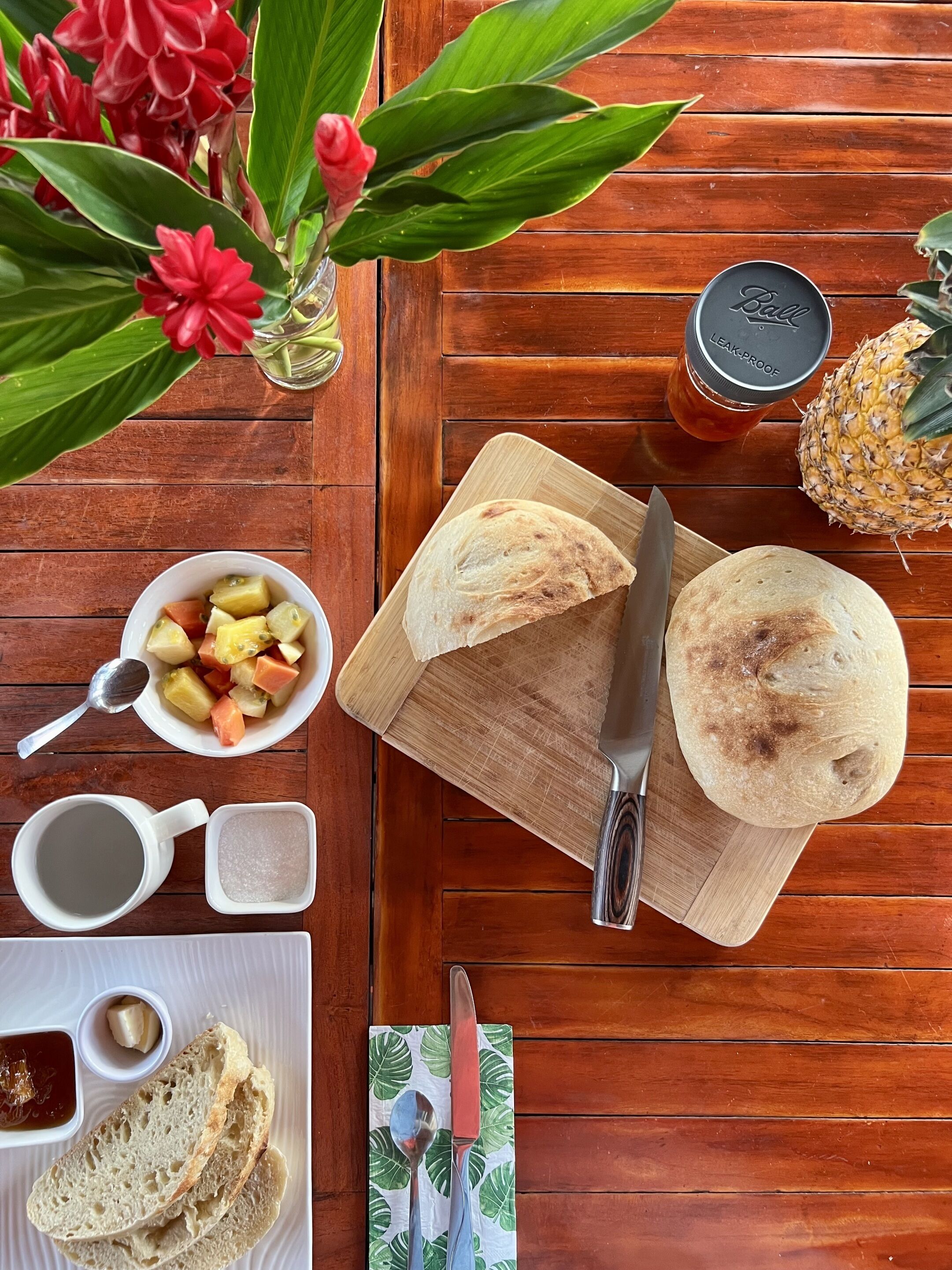 Ein Holztisch mit Schneidebrett, einem Messer, einer Ananas, einer Obstschale, einem Glas Saft, einer Tasse Kaffee, einem Teller mit Brot und einer kleinen Vase mit Blumen.