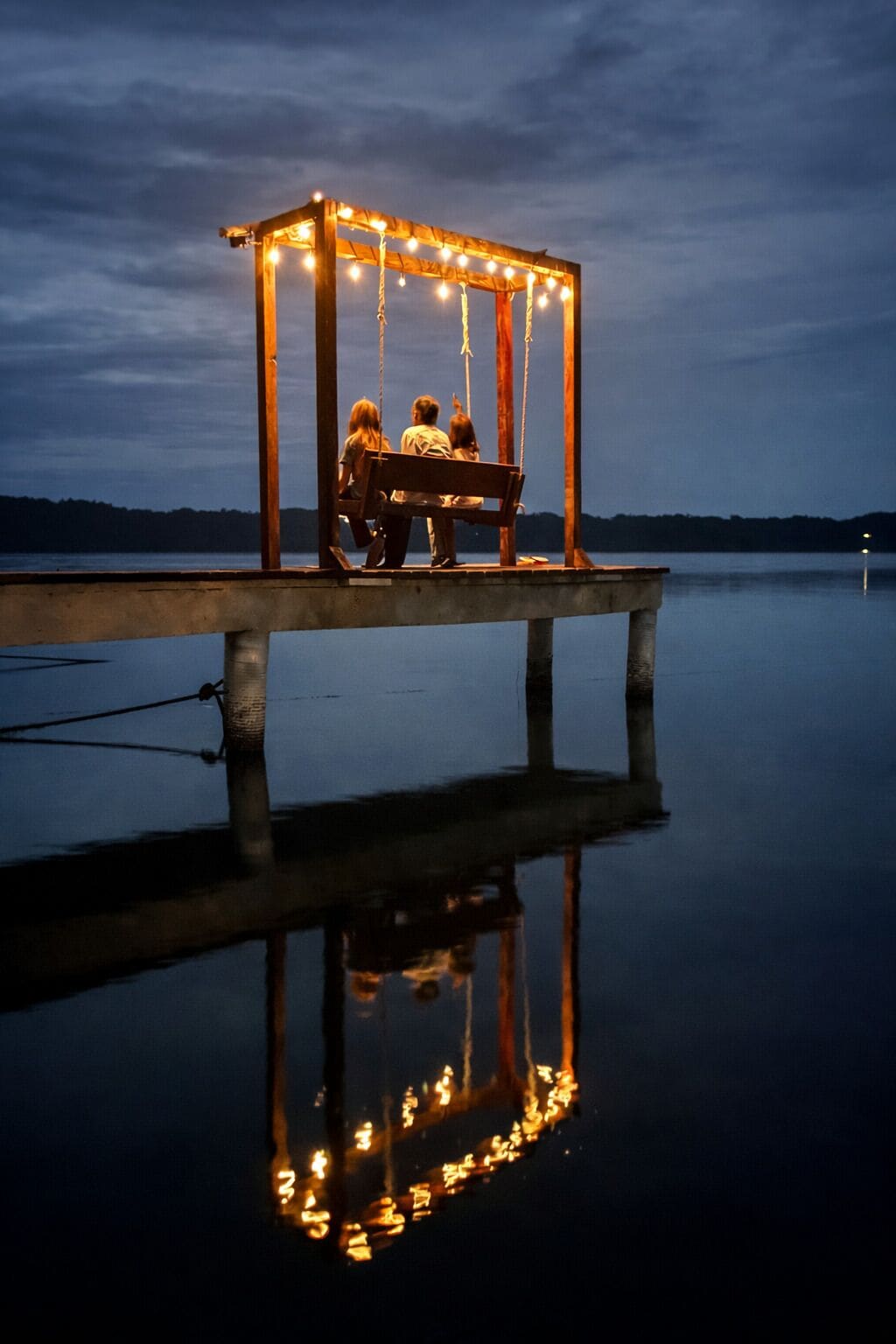 Un belvédère en bois, illuminé de guirlandes lumineuses, avec un banc et une étendue d’eau calme dans laquelle se reflète la structure.