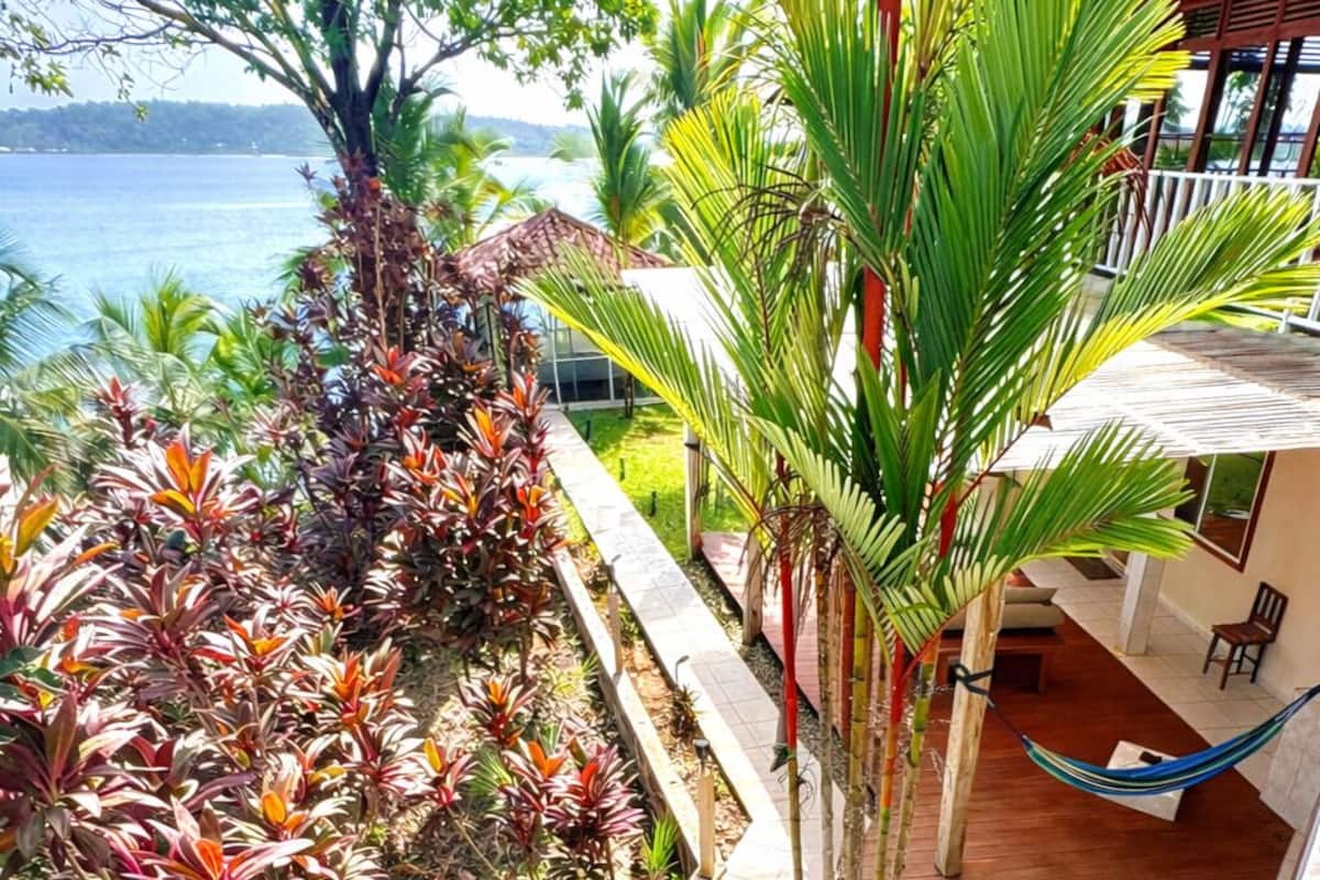 Un jardín tropical con una hamaca, una terraza de madera y vista al mar.