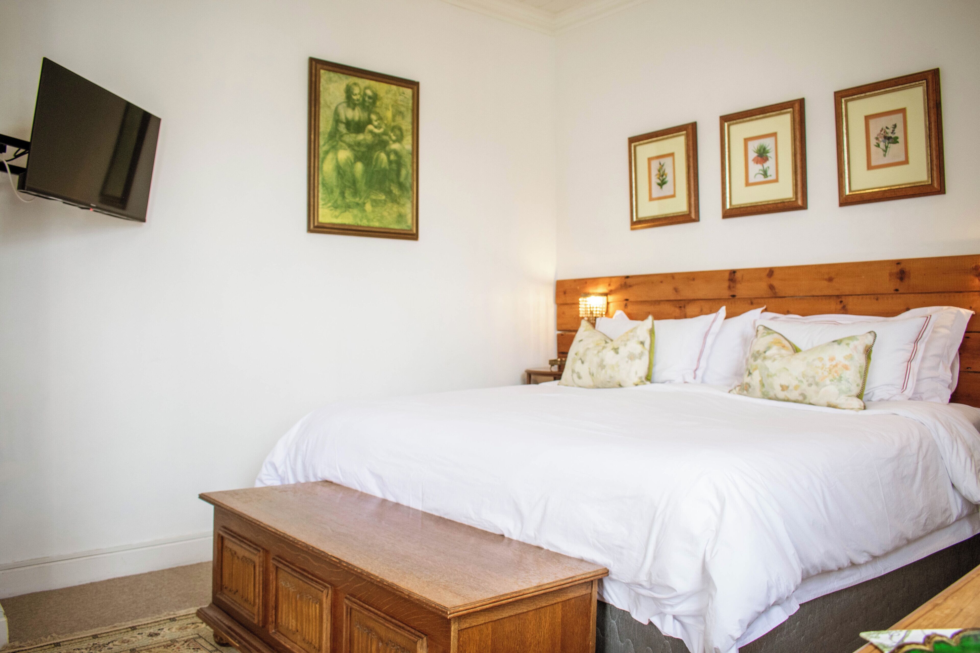 A bedroom with a wooden headboard, a large bed with white bedding, a wooden nightstand, and three framed botanical prints on the wall.