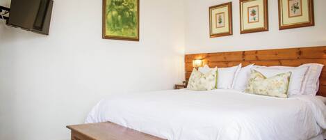 A bedroom with a wooden headboard, a large bed with white bedding, a wooden nightstand, and three framed botanical prints on the wall.