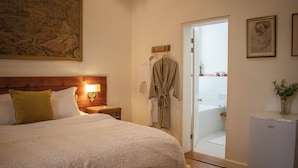 A bedroom with a large bed, a nightstand with a lamp, a wooden headboard, a door leading to a bathroom, and a refrigerator. - Kolonies Tuin (Stellenbosch)