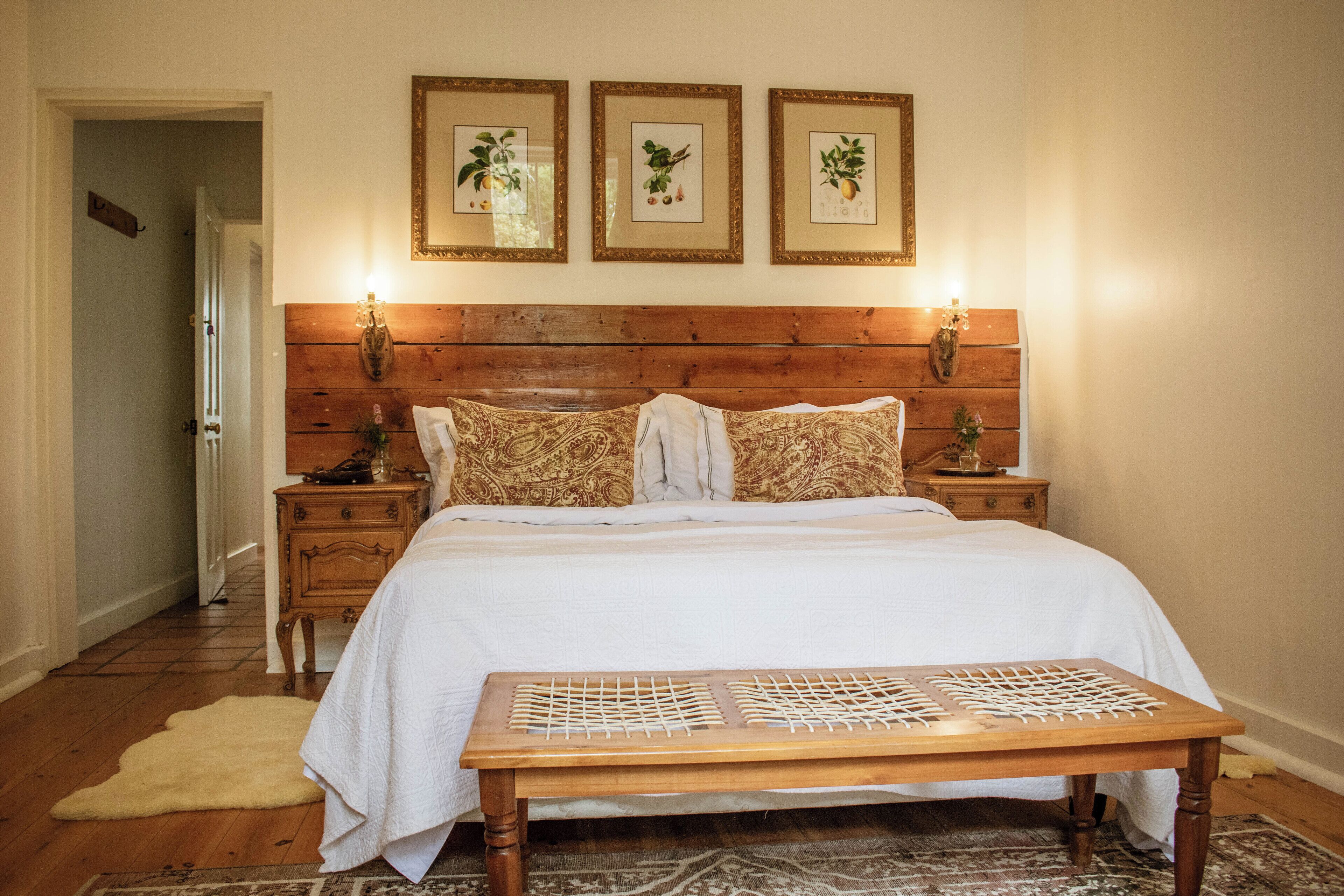 A bedroom with a wooden headboard, a white bedspread, patterned pillows, a bedside table with a lamp, and a wooden bench.