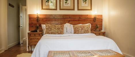 A bedroom with a wooden headboard, a white bedspread, patterned pillows, a bedside table with a lamp, and a wooden bench.