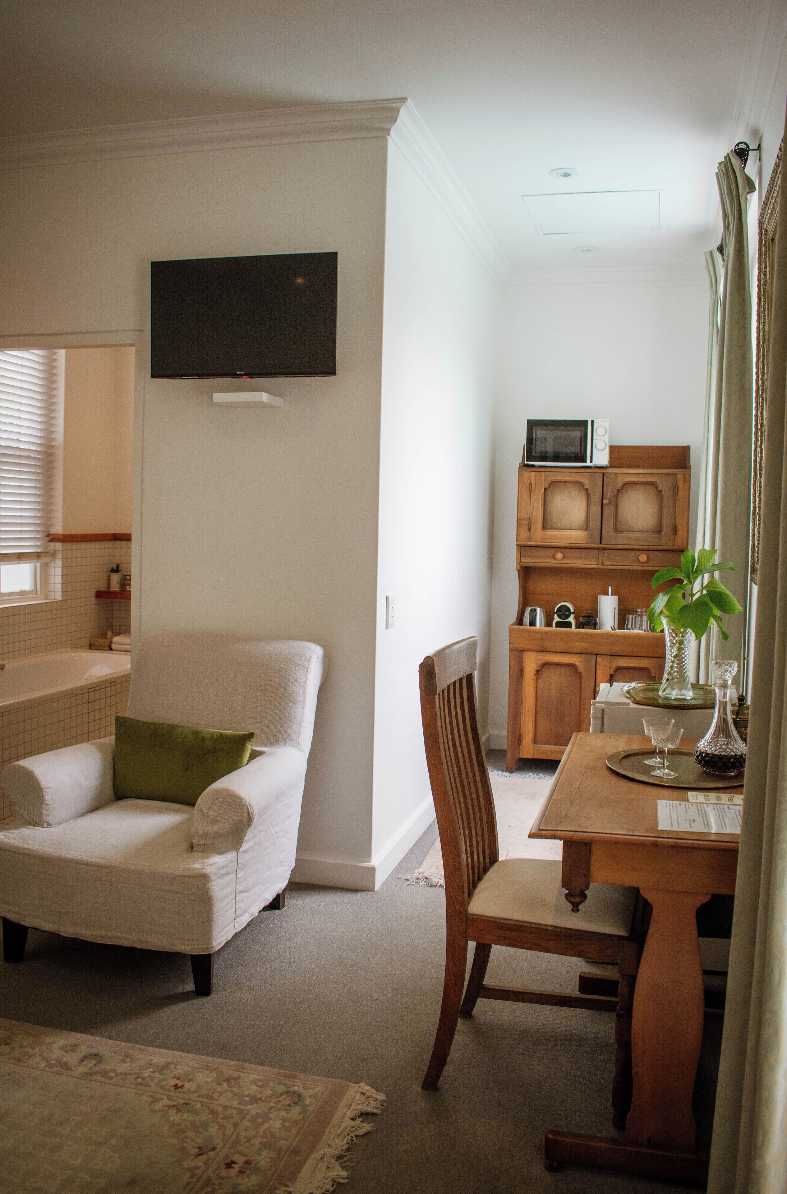 A living room with a white armchair, a wooden dining table with chairs, a flat-screen TV mounted on the wall, and a kitchen area with wooden cabinets and a microwave.