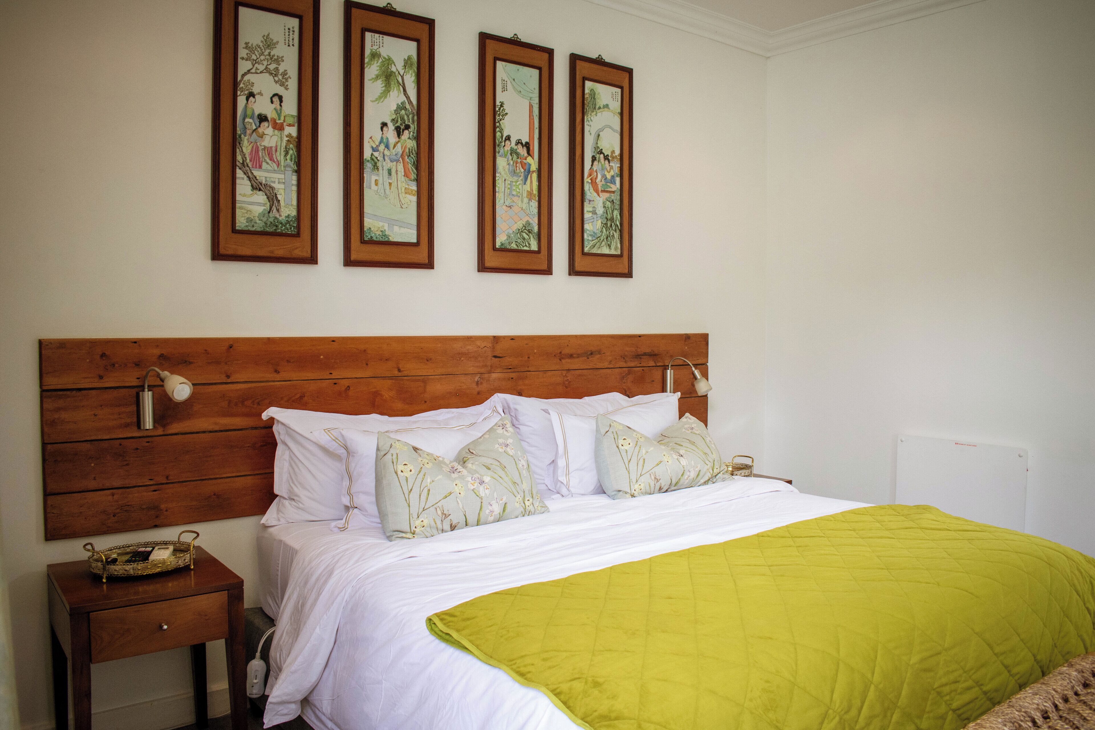 Una cama bien hecha con cabecera de madera, ropa de cama blanca y colcha verde. Encima de la cama hay tres obras de arte enmarcadas, y una mesita de noche con una lámpara y una cesta.