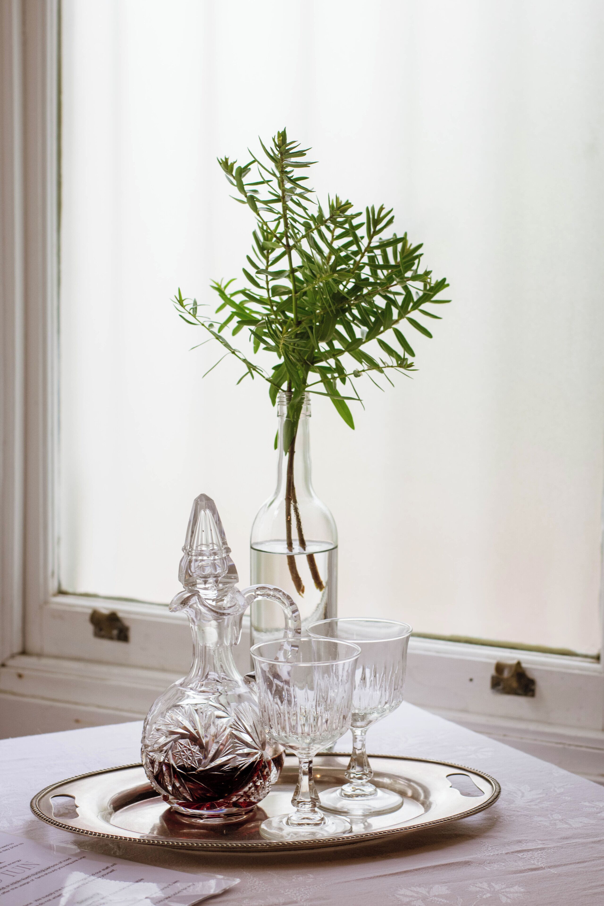 A tray with a crystal decanter, two glasses, and a vase with green foliage on a table by a window.