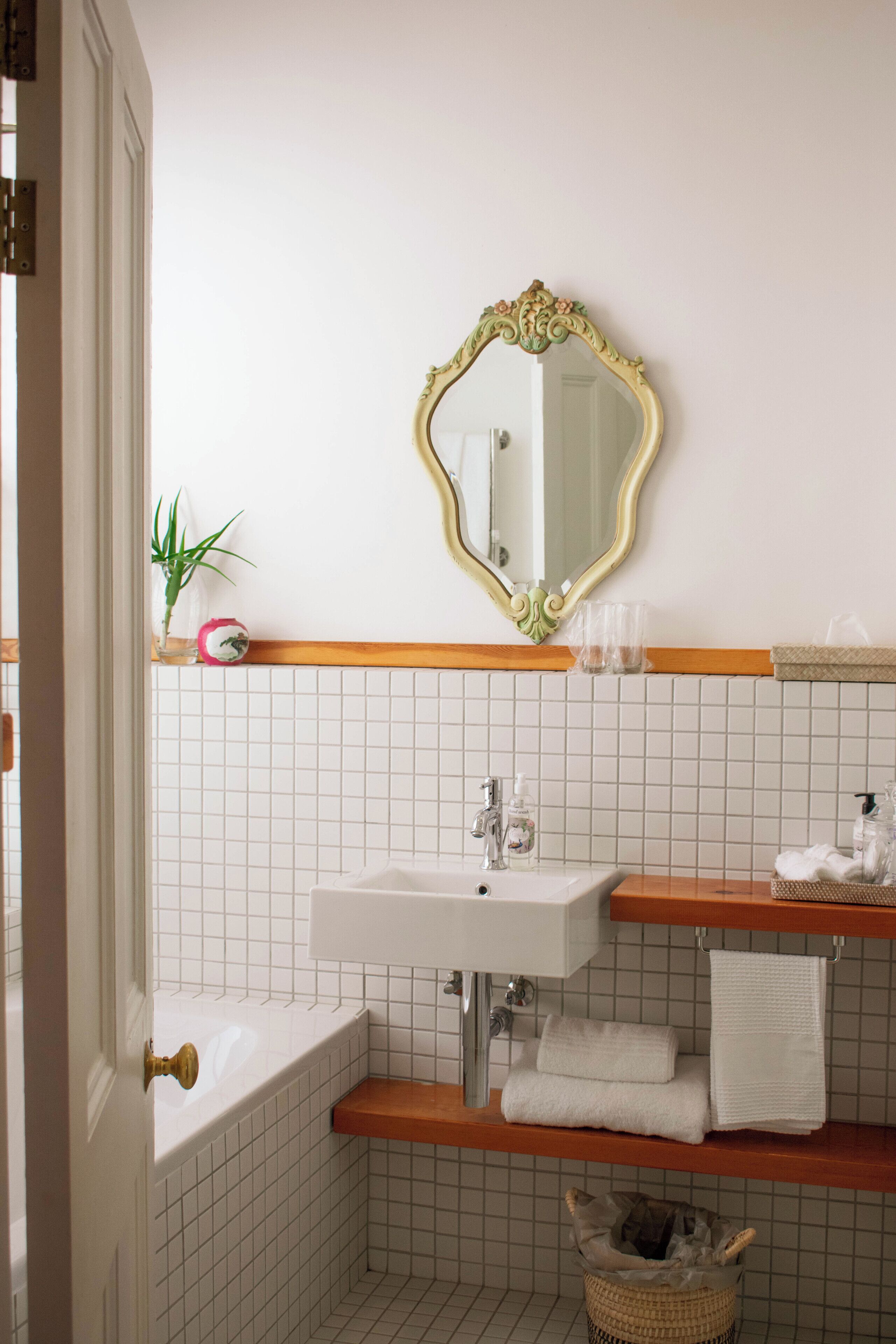 A bathroom with a white sink, a mirror with an ornate frame, a wooden shelf with towels, and a small plant.