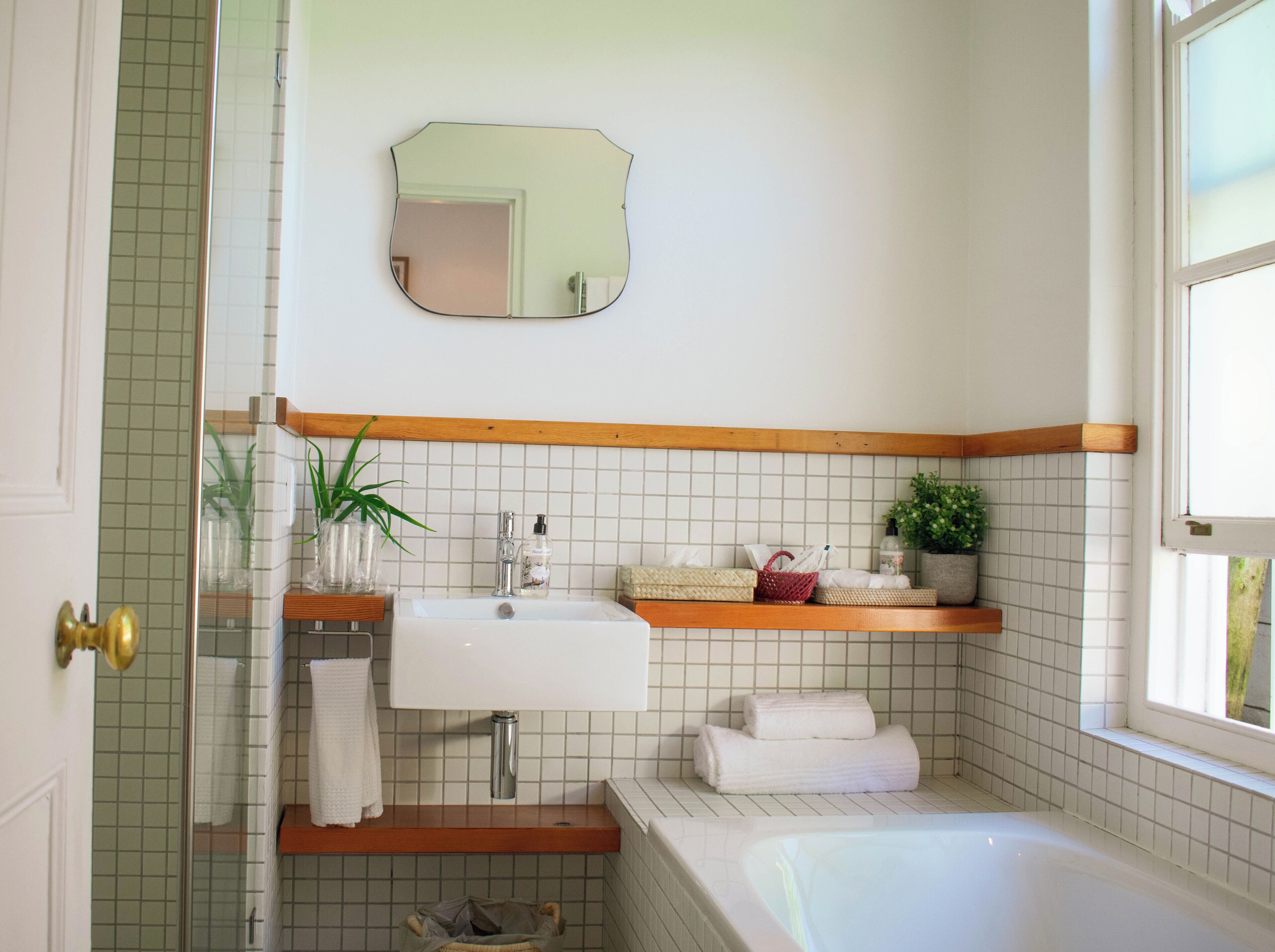 Un baño con lavamanos blanco, repisas de madera, un espejo y una bañera.