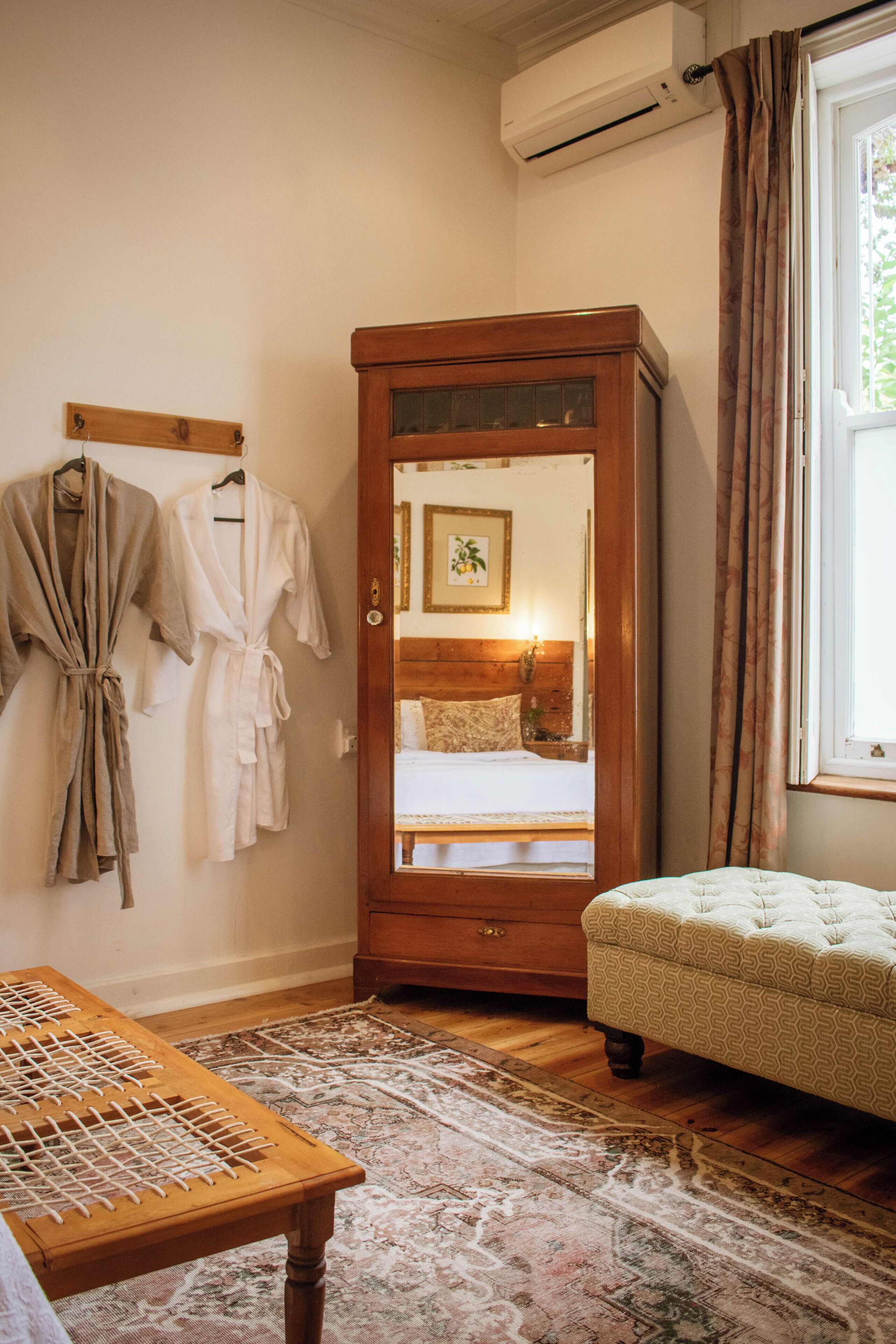 A room with a wooden wardrobe, a bed with a headboard, a small table, and a window with curtains.