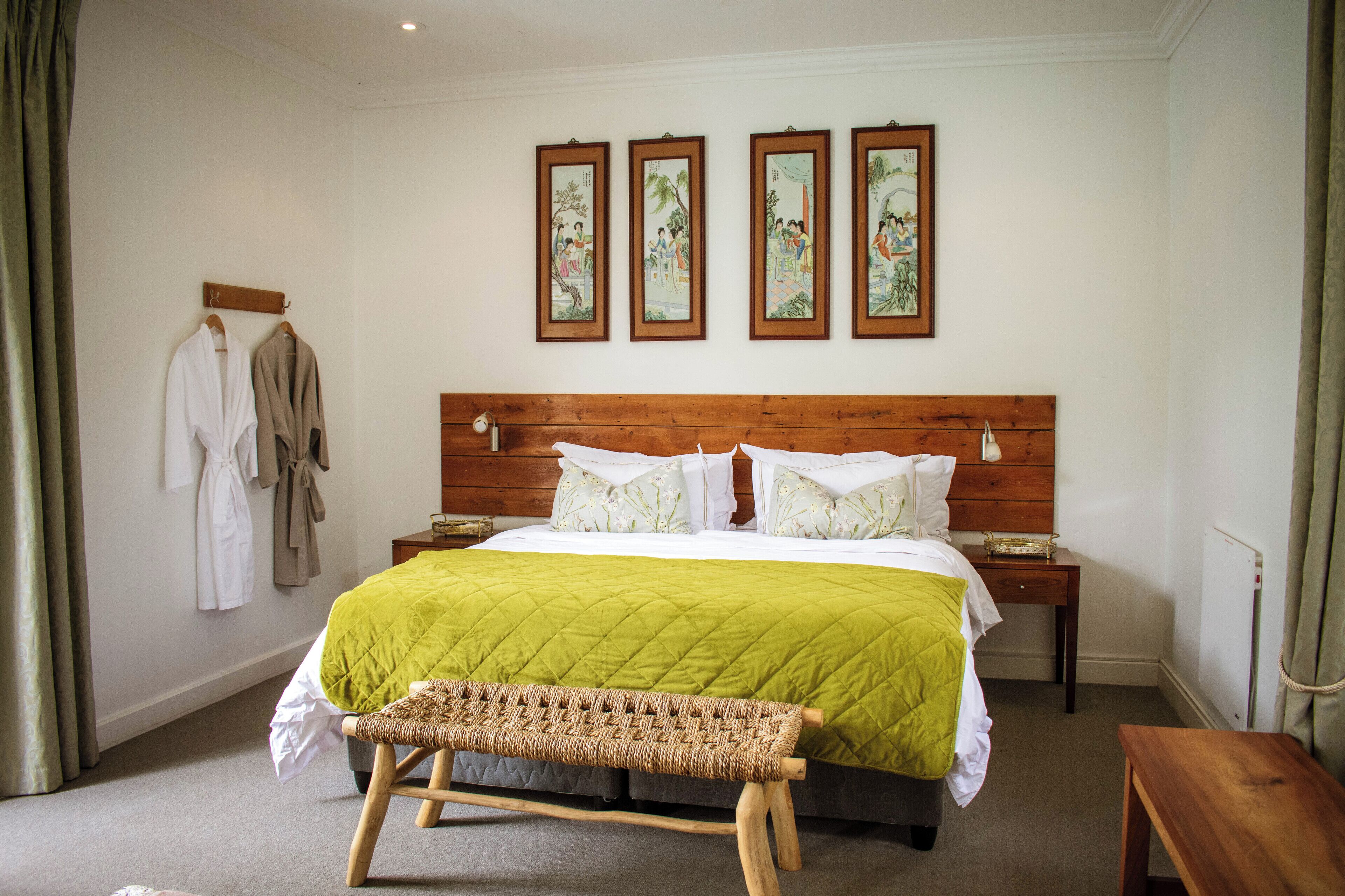 Een slaapkamer met een houten hoofdbord, een groene gewatteerde spreideken, een rieten bankje en drie ingelijste kunstwerken aan de muur.