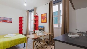 A compact room with a bed, a small dining table, a chair, a red shelf, a window with curtains, and a sink.