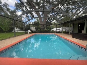 A rectangular swimming pool with a red border, surrounded by a covered patio area with a metal frame and a white chair. - Poolside Family Retreat- Near the Springs & Ocala National Forest. Pet friendly (Ocklawaha)