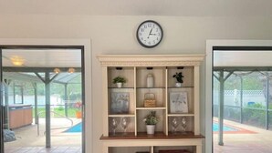 Una habitación con estantería empotrada, un reloj de pared y puerta corredera de cristal que da a un área exterior con piscina.