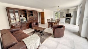 Un vaste salon comprenant un canapé en cuir marron, une table basse et un coin repas avec des chaises et une table en bois. On aperçoit une cuisine équipée d’appareils modernes au fond de la pièce.