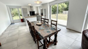 Un vaste espace dédié à la salle à manger, avec une grande table et des chaises, une cuisine en arrière-plan et de grandes fenêtres offrant une vue sur l’extérieur.