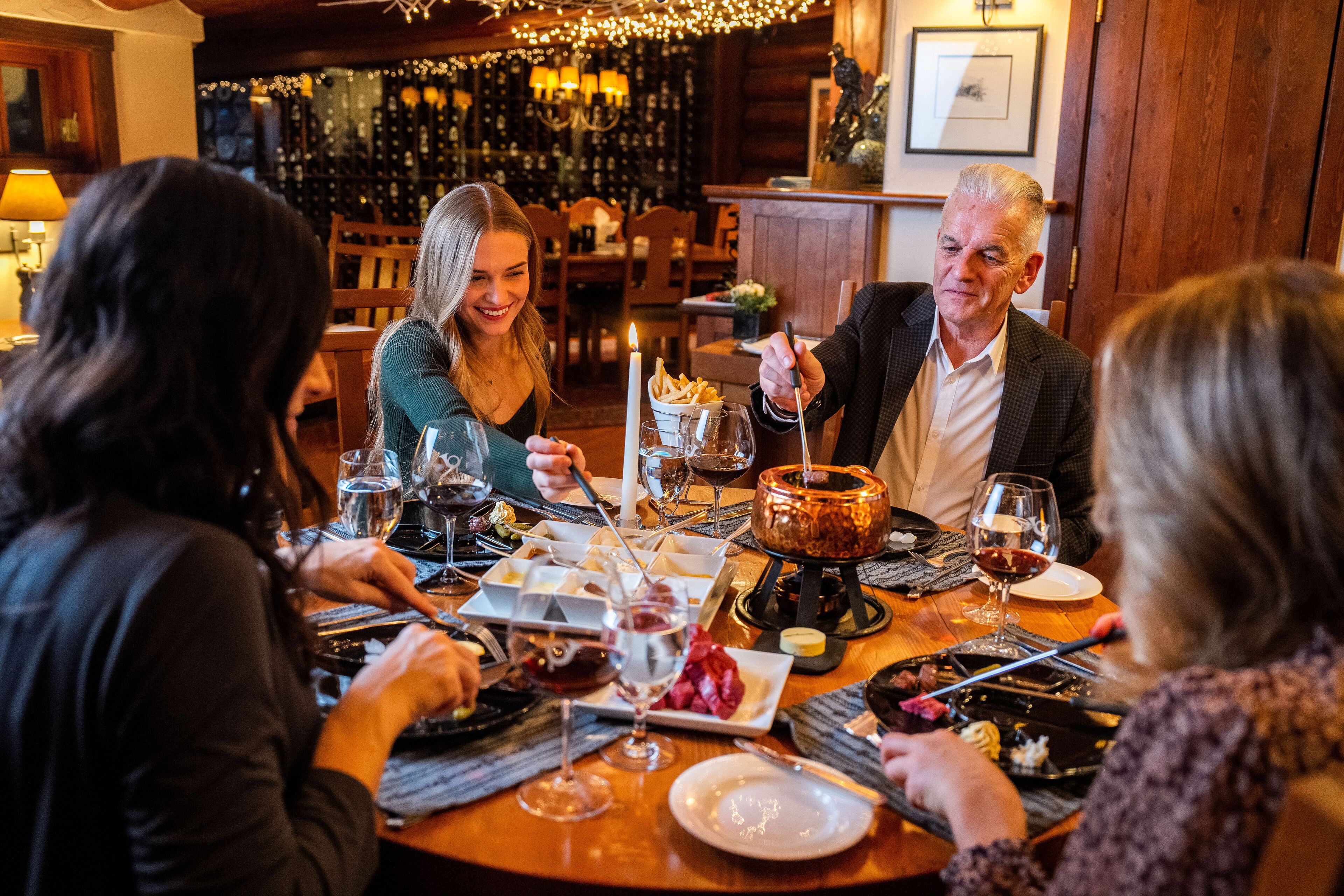 Un groupe de personnes est assis autour d’une table en bois, devant des verres à vin, une bougie et une pierre chaude.