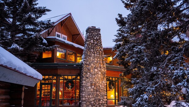 Una cabaña de madera con chimenea de piedra, ventanas amplias y alrededores cubiertos de nieve.
