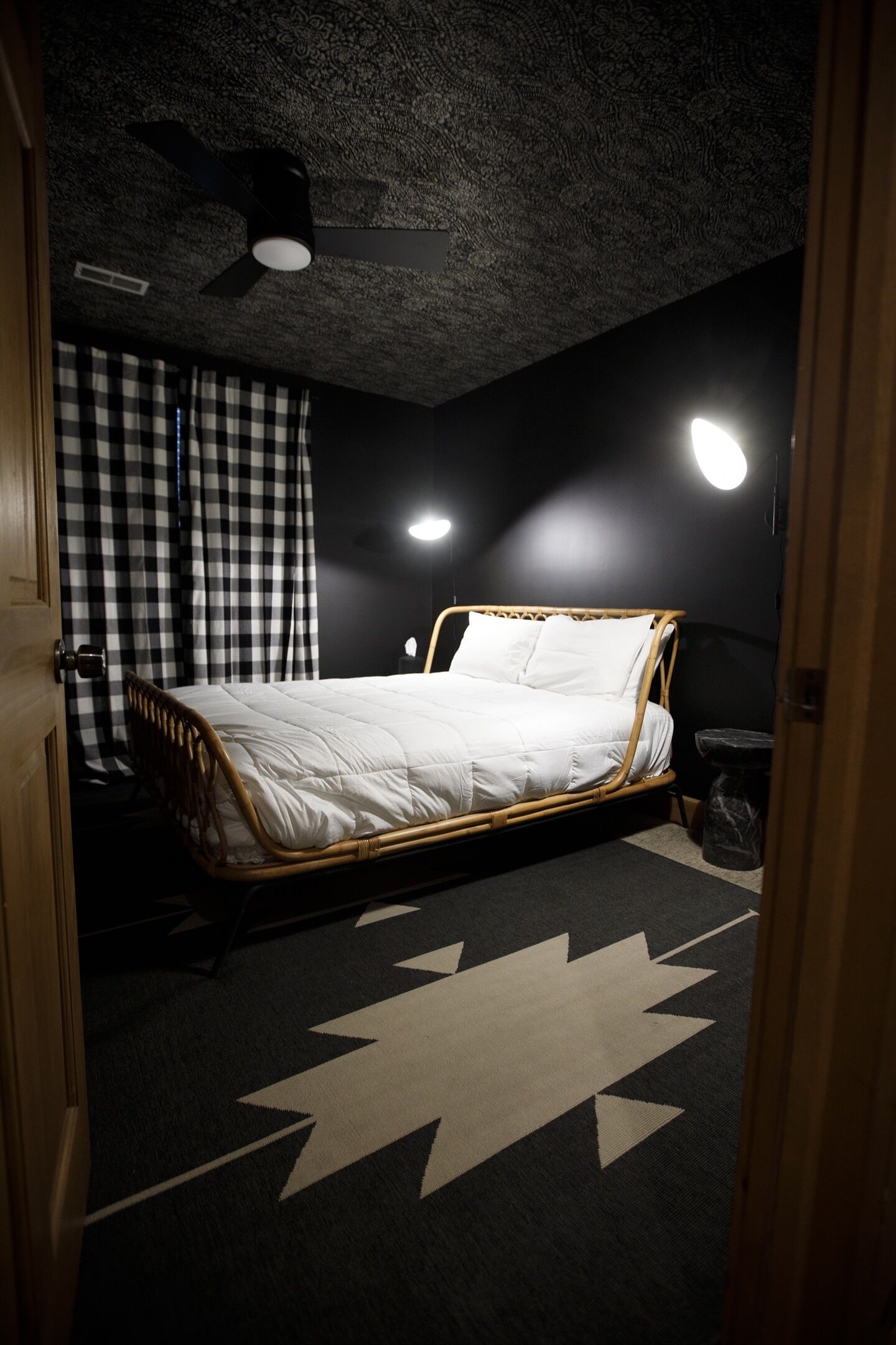 Une chambre à coucher avec un lit, un ventilateur de plafond, une lampe fixée au mur et un rideau à carreaux.