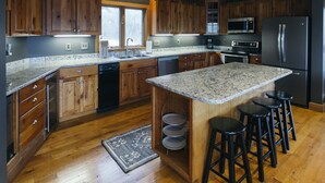A modern kitchen with wooden cabinets, granite countertops, a stainless steel refrigerator, and a built-in island with bar stools. - Among the Trees - The Lodge (Logan)