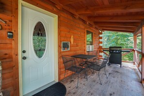 A wooden cabin with a white door, a small porch with a table and chairs, and a grill. - Sandstone Oasis | Hocking Hills, Couples, Hot Tub (Logan)
