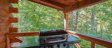 A wooden deck with a charcoal grill and a table, surrounded by a forested area.