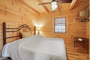 Dormitorio de una cabaña de madera con cama, mesita de noche, lámpara, ventana con persianas y ventilador de techo.