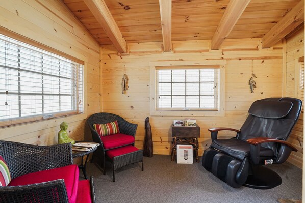 A wooden cabin interior with a wicker chair, a massage chair, and a small table with books. - Sandstone Oasis | Hocking Hills, Couples, Hot Tub (Logan)