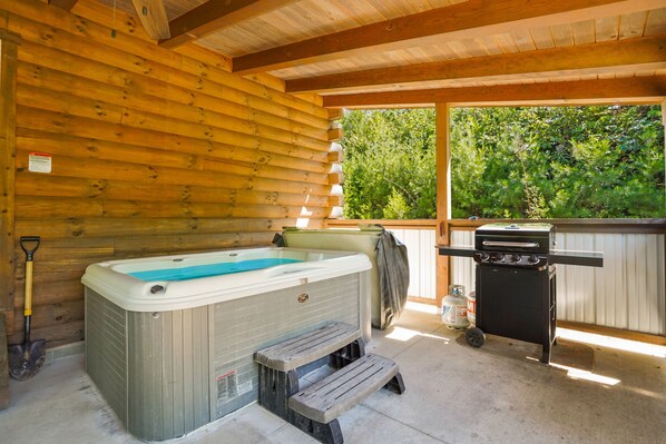 A wooden cabin with a hot tub, a grill, and a window overlooking greenery.