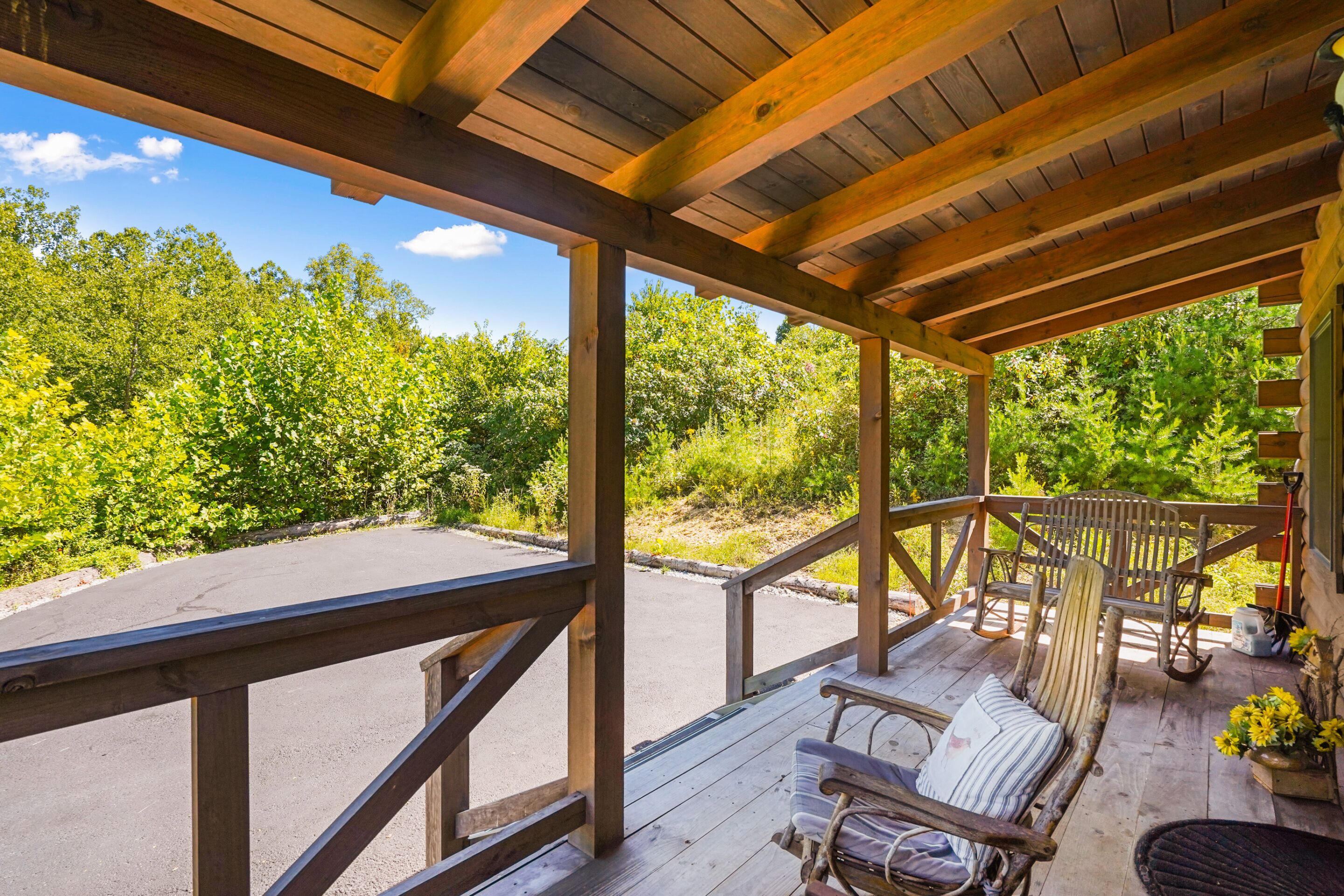 A wooden deck with a bench and a chair, overlooking a parking area and dense greenery.
