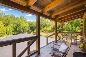 A wooden deck with a bench and a chair, overlooking a parking area and dense greenery. - Lovebirds Landing | Hocking Hills, Couples Cabin (Logan)