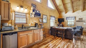 A wooden cabin interior with a kitchen, dining area, and a large window. - Lovebirds Landing | Hocking Hills, Couples Cabin (Logan)