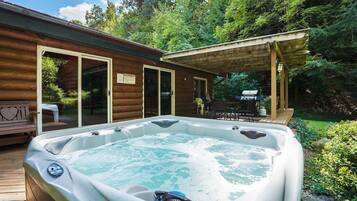 Un jacuzzi en una cabaña de madera con un porche cubierto y una parrilla afuera.