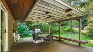 Une terrasse couverte avec une table et des chaises en bois, un barbecue et une vue sur un jardin luxuriant.