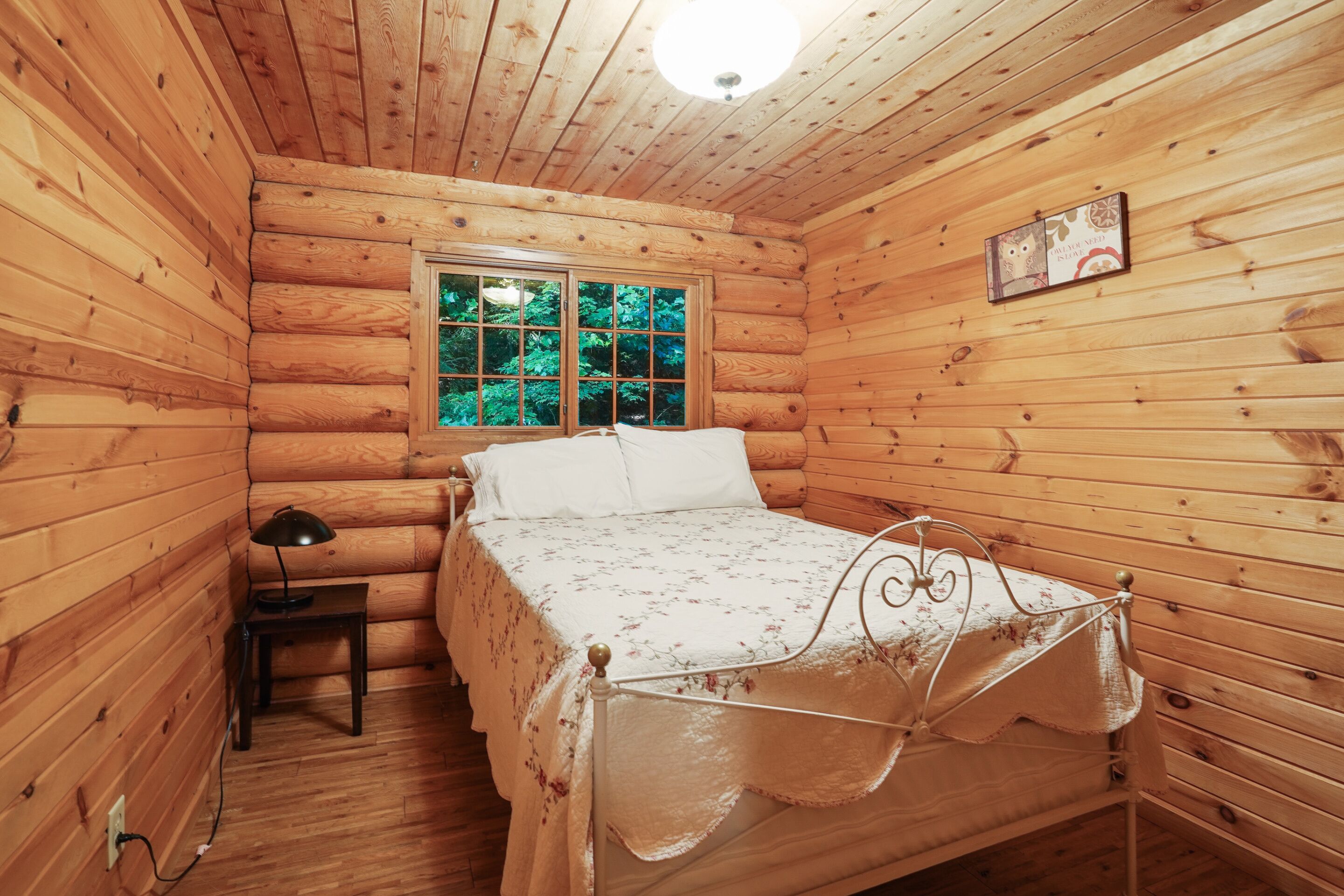 Ein Schlafzimmer in einer Holzhütte mit einem Bett, einem Nachttisch, einer Lampe und einem Fenster mit Blick auf Grünflächen.