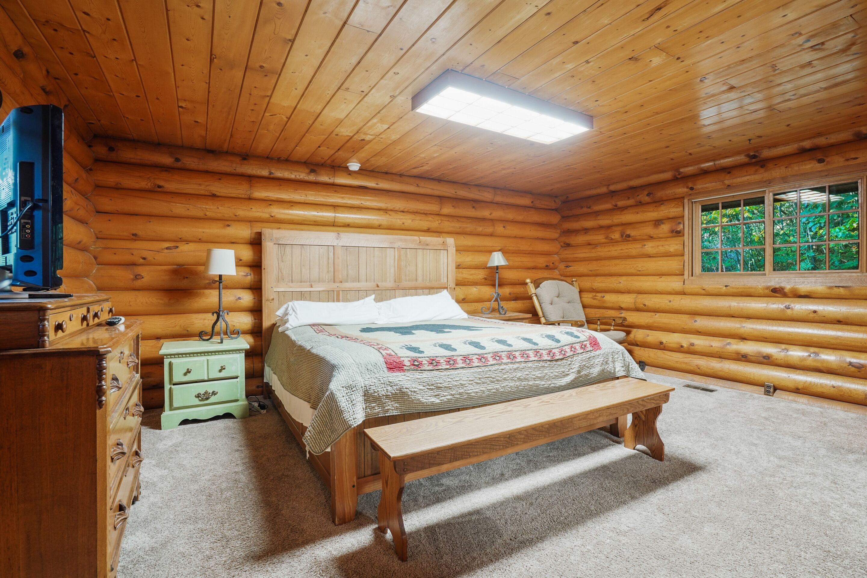 Un dormitorio tipo cabaña de madera con una cama de madera, lámparas de noche, un banco, un aparador y una ventana con vistas a zonas verdes.