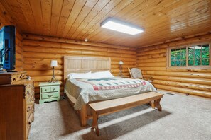 A log cabin bedroom with a wooden bed, bedside lamps, a bench, a dresser, and a window with a view of greenery.