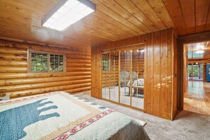 Et interiør af en hytte med en stor seng, en skydedør, der fører til en balkon, og et vindue med udsigt til grøn bevoksning.