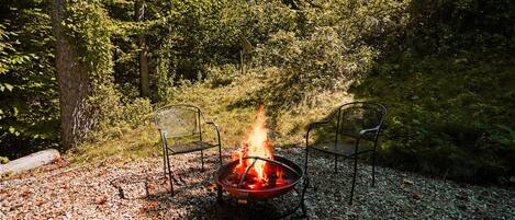 Een vuurkorf met twee stoelen, omgeven door grind en bomen.