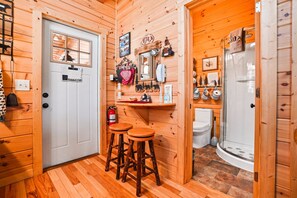 A wooden cabin interior with a door, two stools, a mirror, a shelf with various items, a toilet, and a shower area. - Snuggle Bear | Hocking Hills, Adorable Tiny Home (Logan)